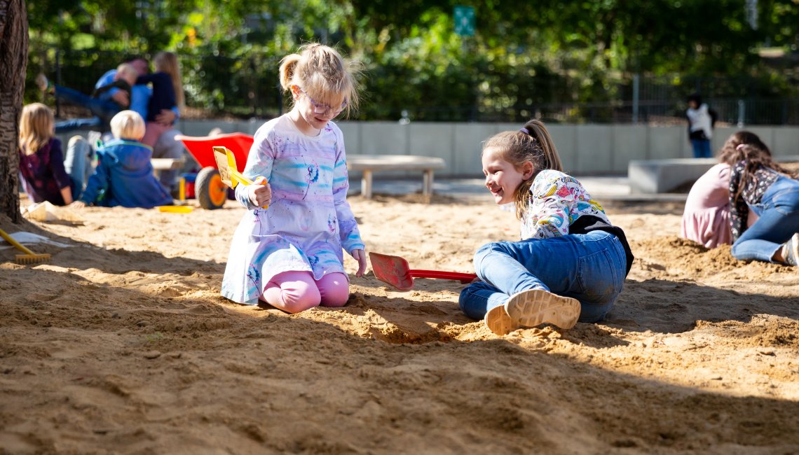 Zwei M&auml;dchen spielend lachend in einem Sandkaste. Im Hintergrund spielen weitere Kinder.